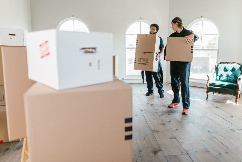 Zwei Mitarbeiter tragen beschriftete Umzugskartons durch eine helle, leere Wohnung